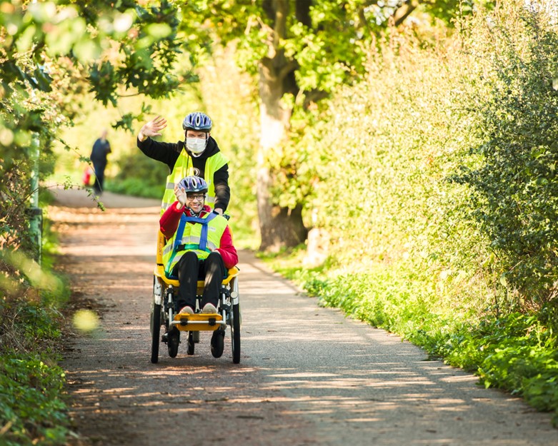 An image containing two adult males on an adapted bicycle in a leafy lane near Lowestoft Community Hub. Both are wearing high vis jackets and cycle helmets and waving at the camera. The male support worker is also wearing a mask to reduce Covid Risks.