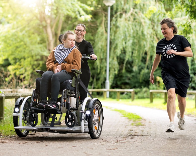 An image containing an adult female customer on an adapted bicycle ridden by a female support worker while another female support worker runs alongside, they are riding in the leafy grounds of Stowmarket Community Hub. Both the support workers are smiling and the customer is interacting with them